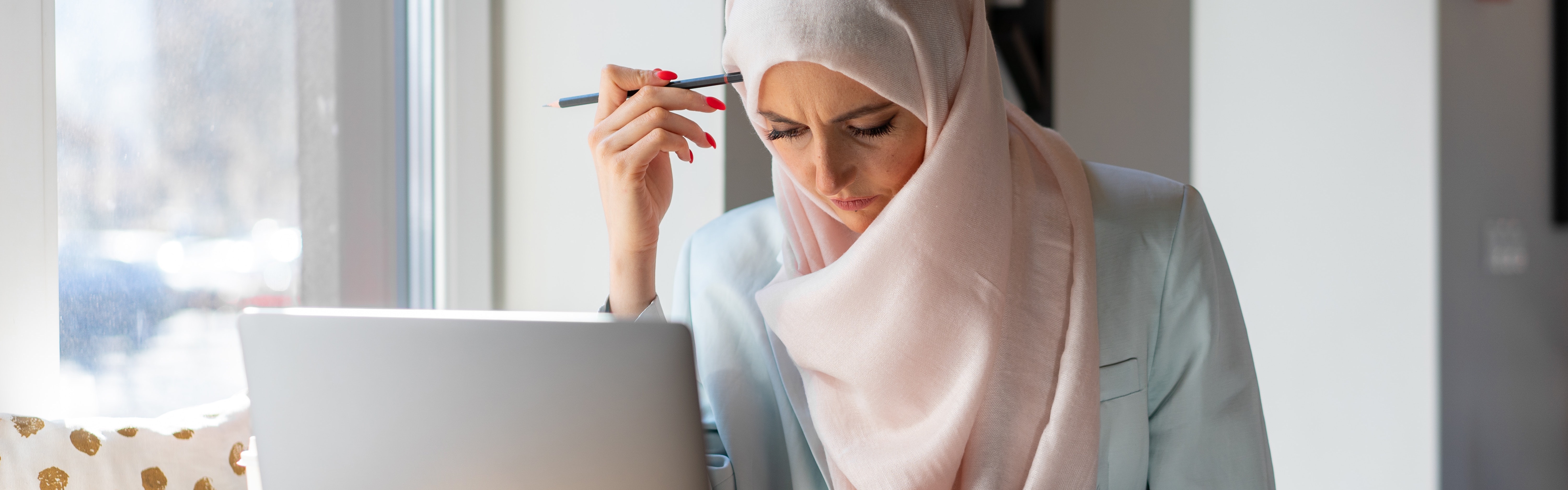 A teacher prep candidate sits at her laptop studying for her Missouri certification exams. She is wearing a hijab and holding a pencil against her head while she reads. 