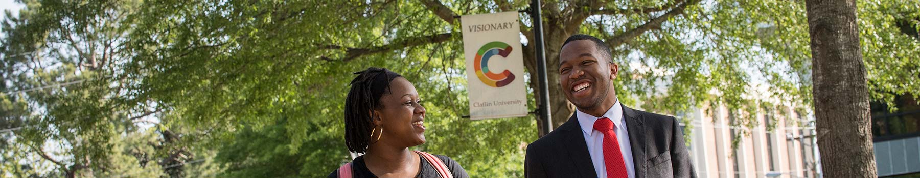 A man and a woman of color, smiling under the shade of trees in the Claflin University